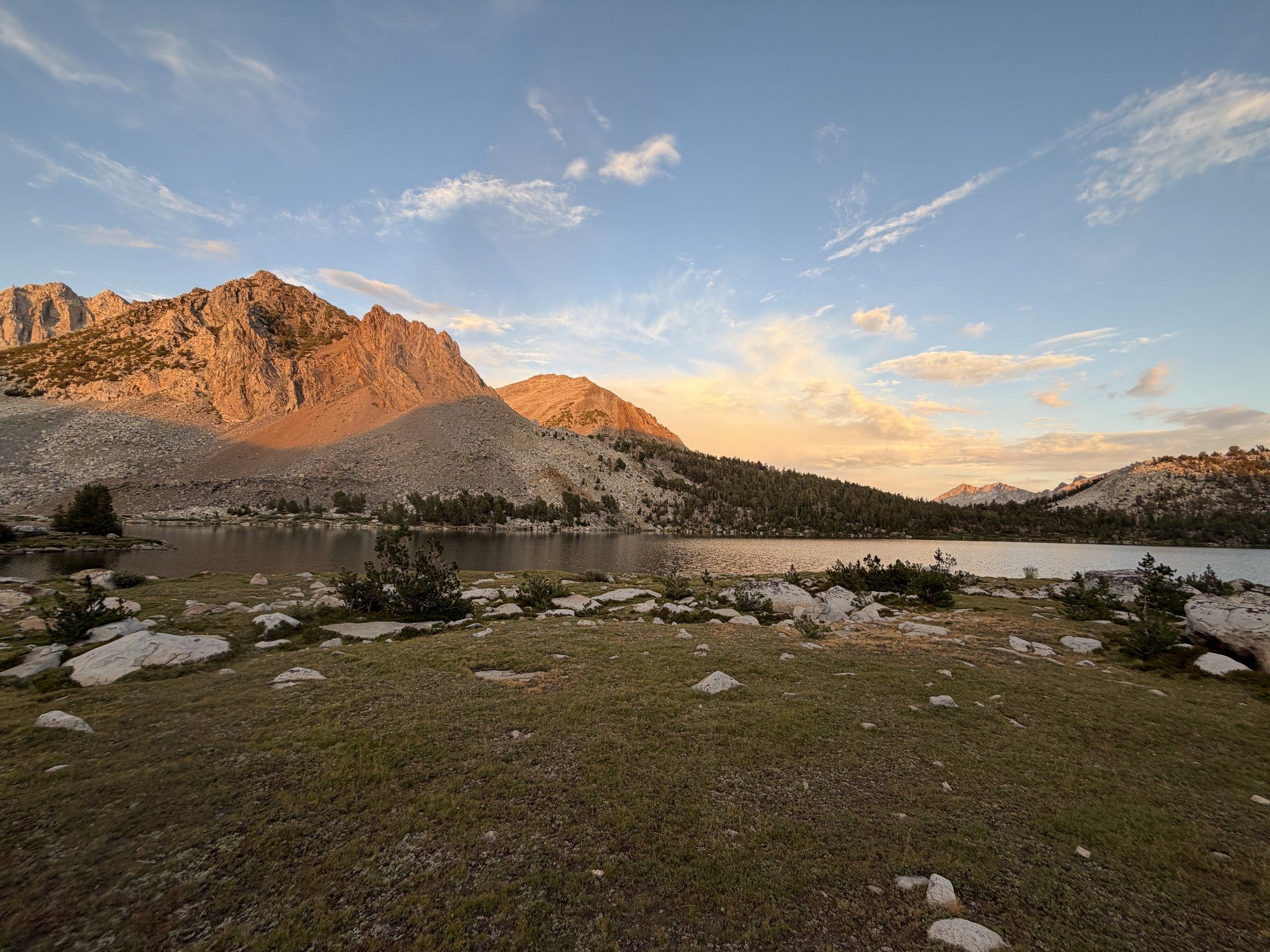 Warm light on a ridge above a still lake, with meadow and scattered granite in the foreground